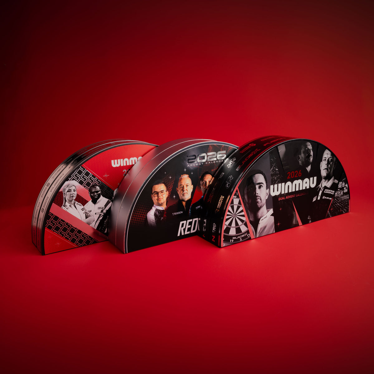 Three Winmau and Red Dragon Darts semi-circular darts advent calendars arranged in a row on a red studio background. The calendars feature black and red graphics with player portraits and the text 'winmau' and 'red dragon'.