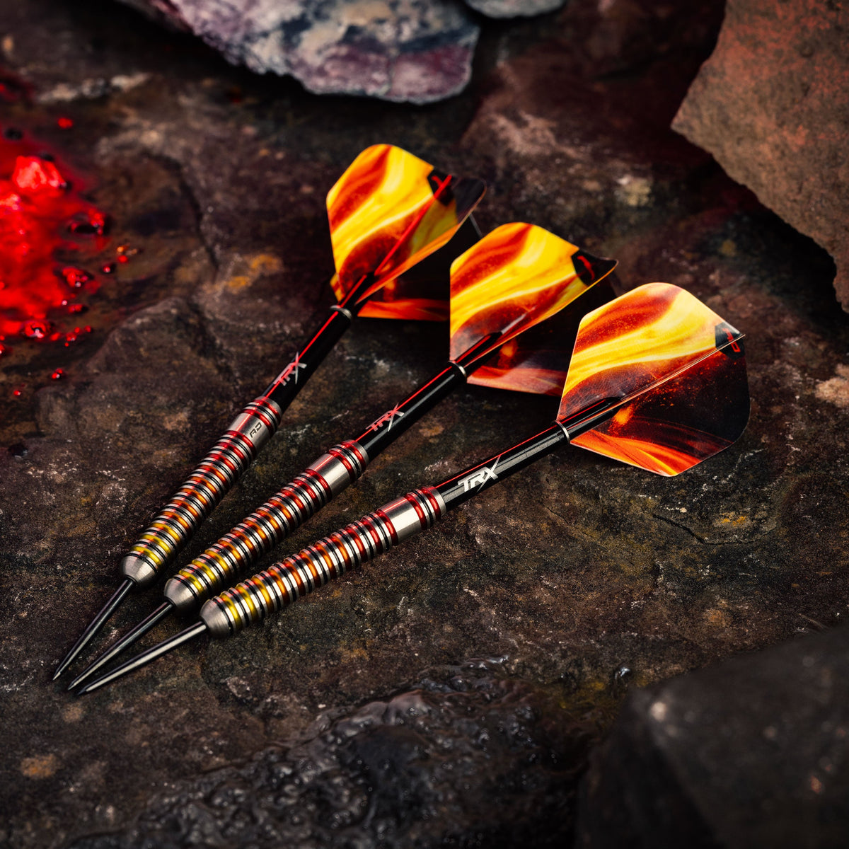 Three Red Dragon darts with metallic gold and red striped handles and yellow-orange patterned flights resting on a dark textured stone surface.