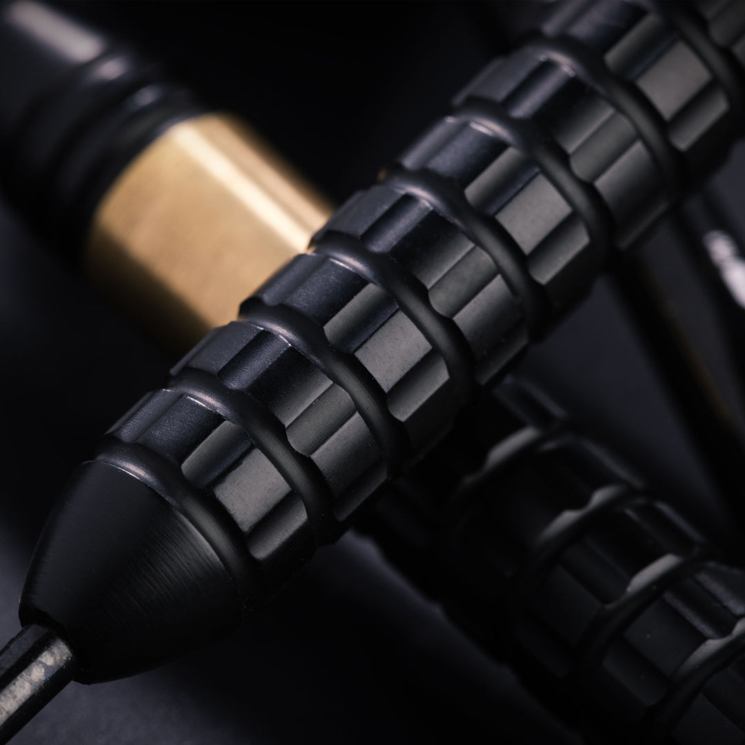 Close-up of a black textured dart grip with a metallic gold section in the background, set against a dark backdrop.