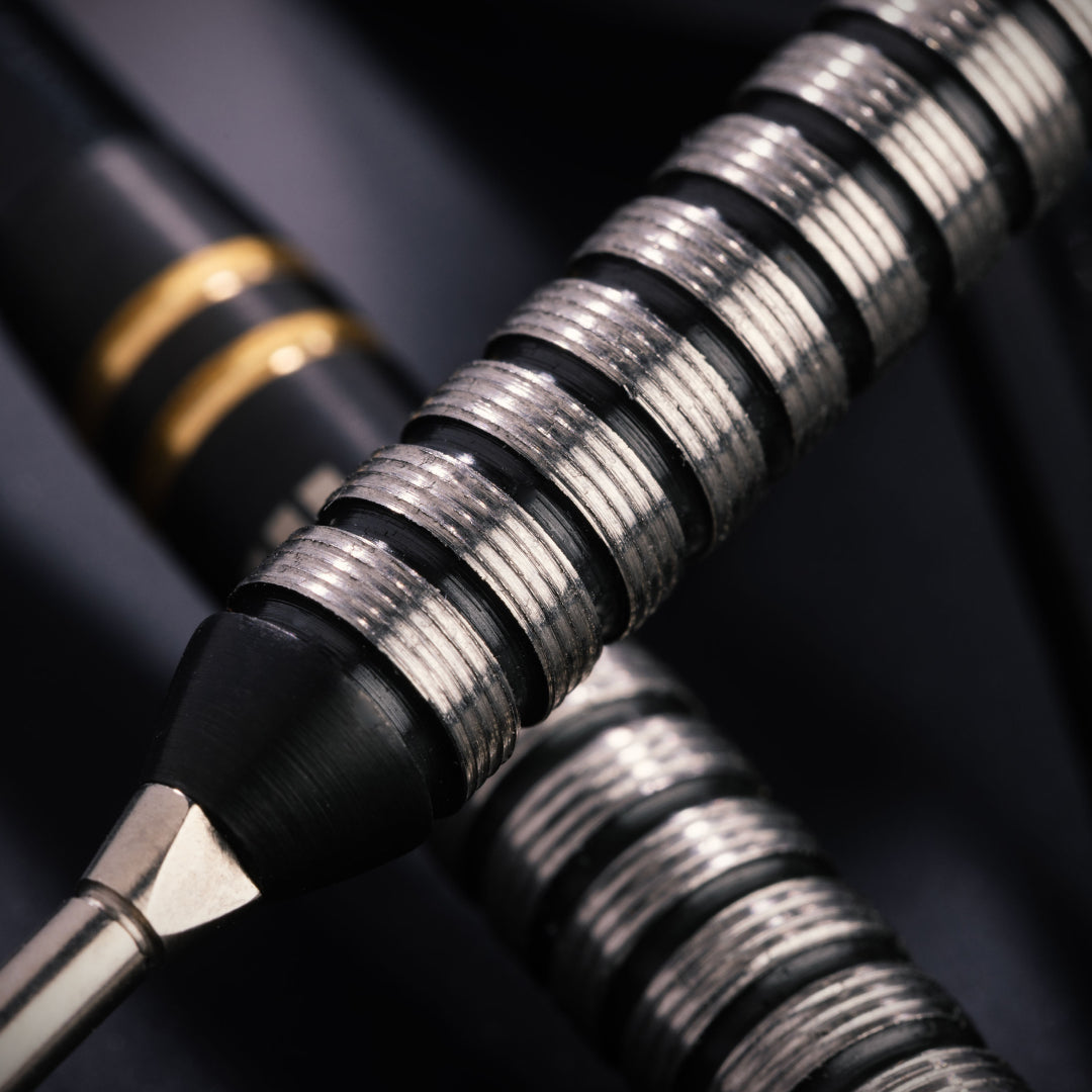Close-up of a black and silver dart with a textured metallic grip, shown against a dark background with another dart partially visible.